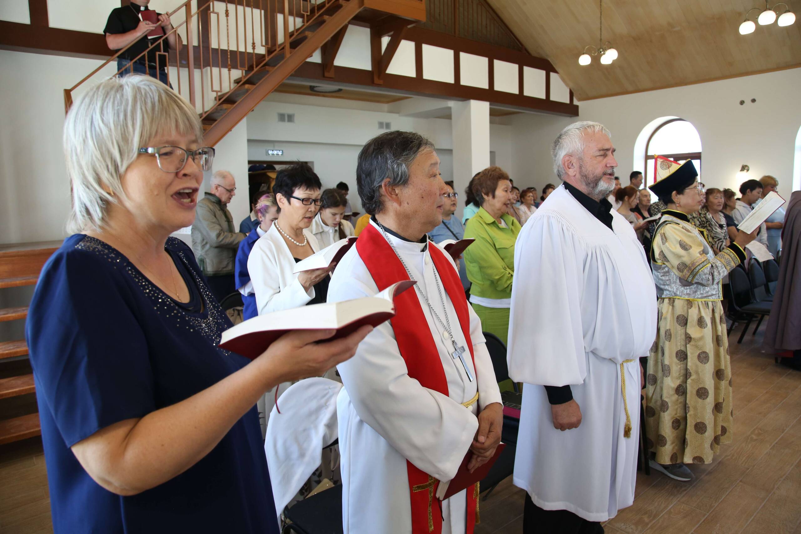 Ihmiset laulavat seisaaltaan. Etualalla nainen ja hänen vierellään kaksi papin albaan pukeutunutta miestä. Kirkkosali on vaalea ja valoisa.