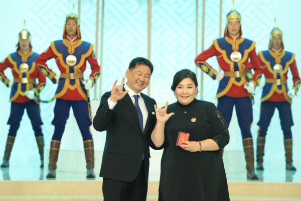 A man and a woman smiling and doing sing language sings, soldier in background