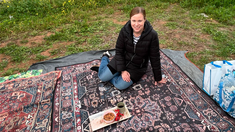 A woman sitting on a carpet outside in the nature, serving tea.